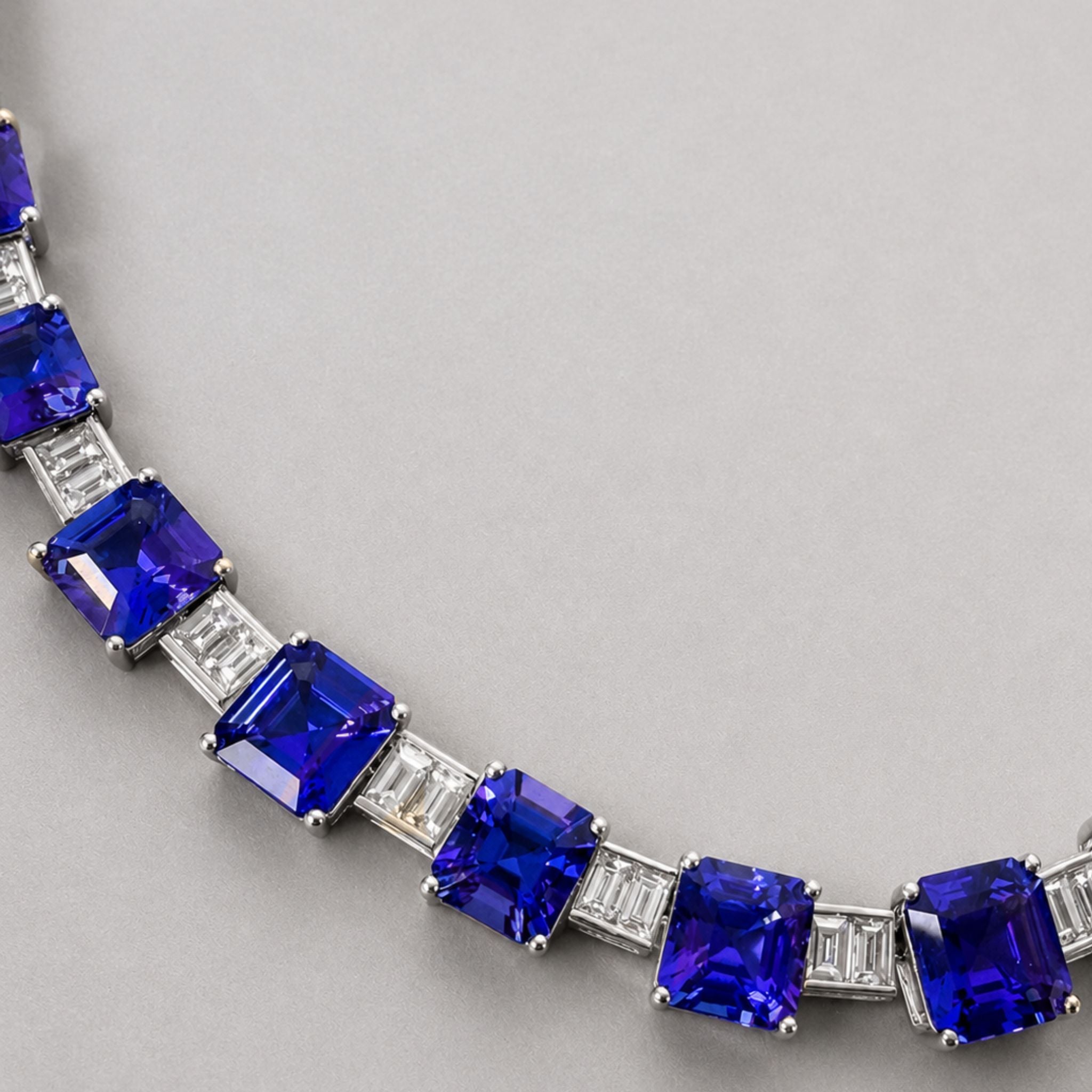 A close-up of necklace featuring large, deep blue emerald-cut tanzanites  set with baguette-cut diamonds stones, photographed on a neutral gray background.